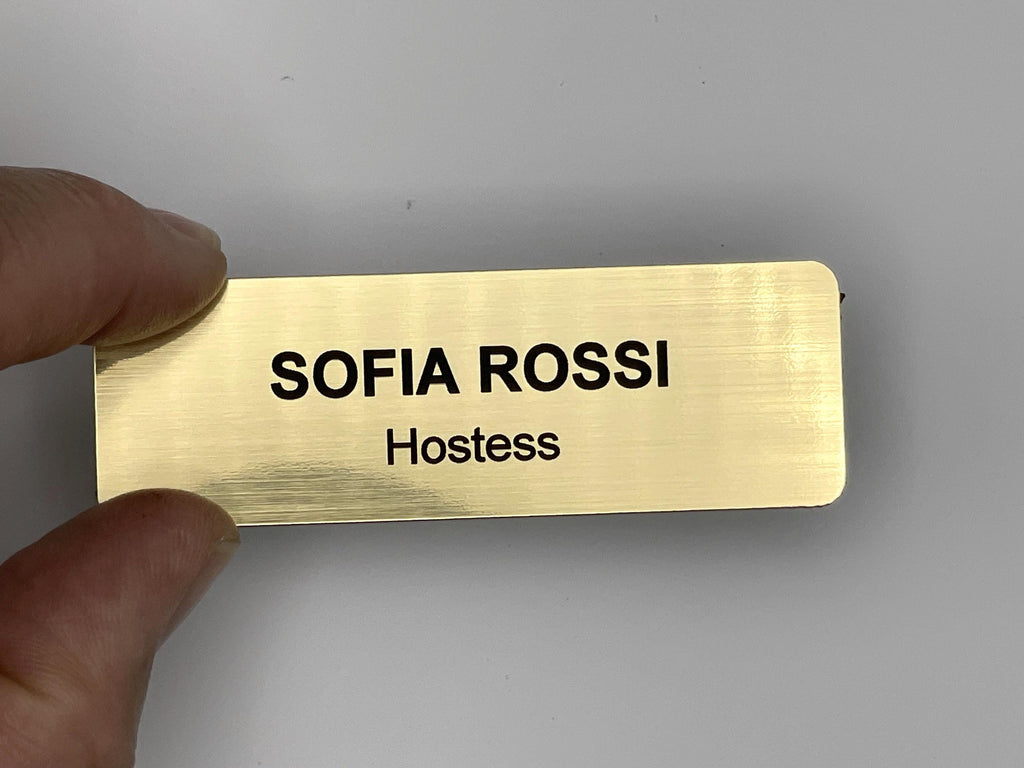A rectangular gold-colored name badge with a brushed metal-look finish. The text SOFIA ROSSI is engraved in black capital letters on the top line, and the word Hostess is engraved in black letters on the line below.