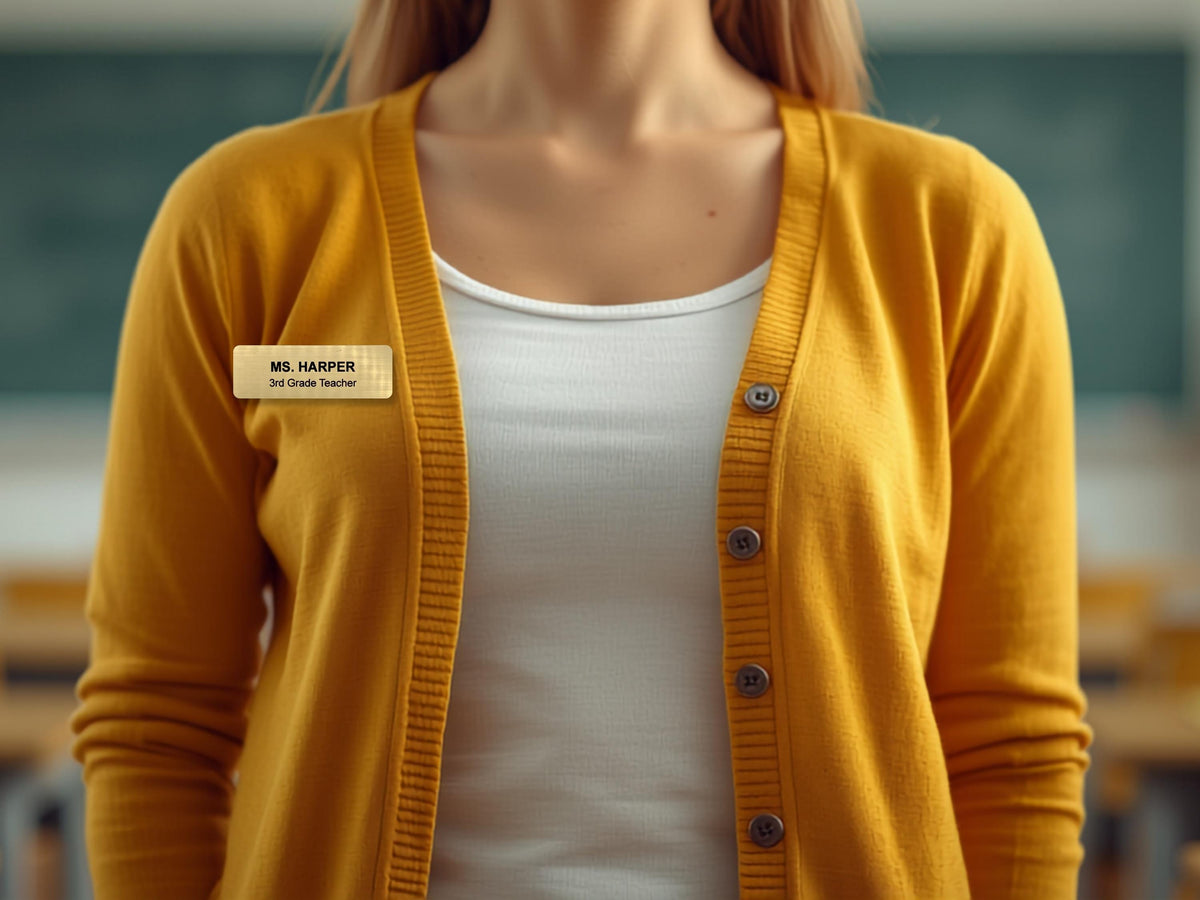 A mustard yellow button-down cardigan worn over a white scoop-neck top. A rectangular gold-toned name tag is pinned to the left side of the cardigan, featuring the black text MS. HARPER and the smaller text 3rd Grade Teacher below it.