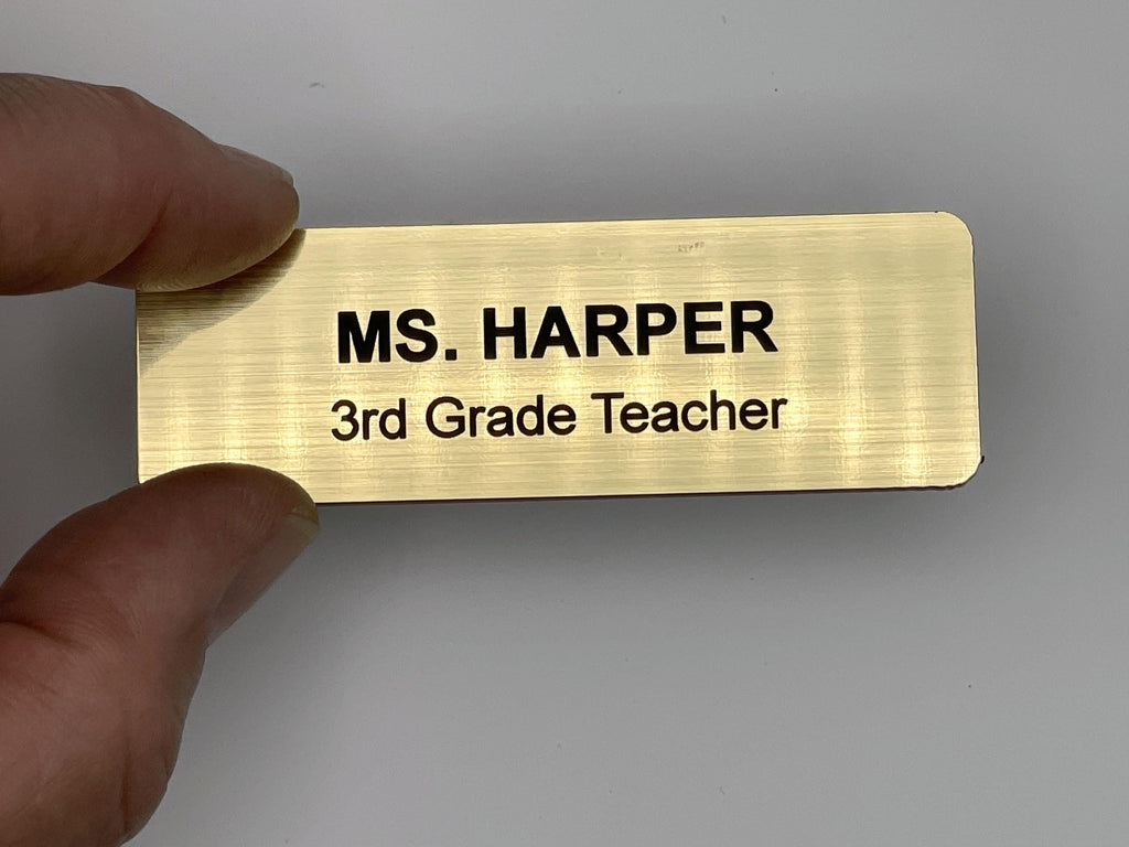 A rectangular gold-toned name badge with a brushed metal-look finish. The text is engraved in black, reading MS. HARPER on the top line and 3rd Grade Teacher on the bottom line. The badge has rounded corners and a smooth, metal-look surface.
