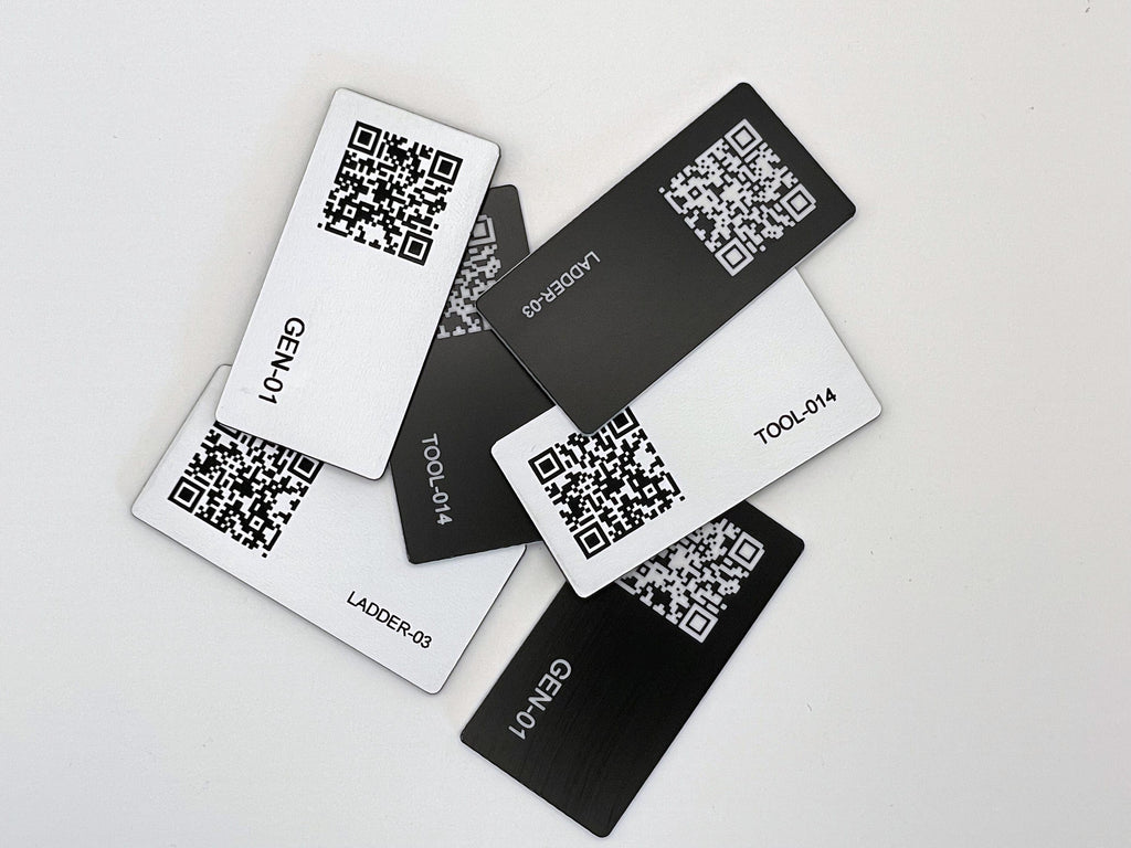 A collection of rectangular engraved tags in black and white, each featuring a unique QR code and engraved text. The labels include identifiers such as GEN-01, LADDER-03, and TOOL-014, arranged in a scattered overlapping pattern.