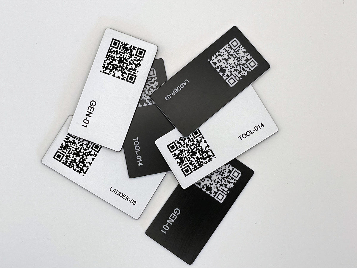 A collection of rectangular engraved tags in black and white, each featuring a unique QR code and engraved text. The labels include identifiers such as GEN-01, LADDER-03, and TOOL-014, arranged in a scattered overlapping pattern.