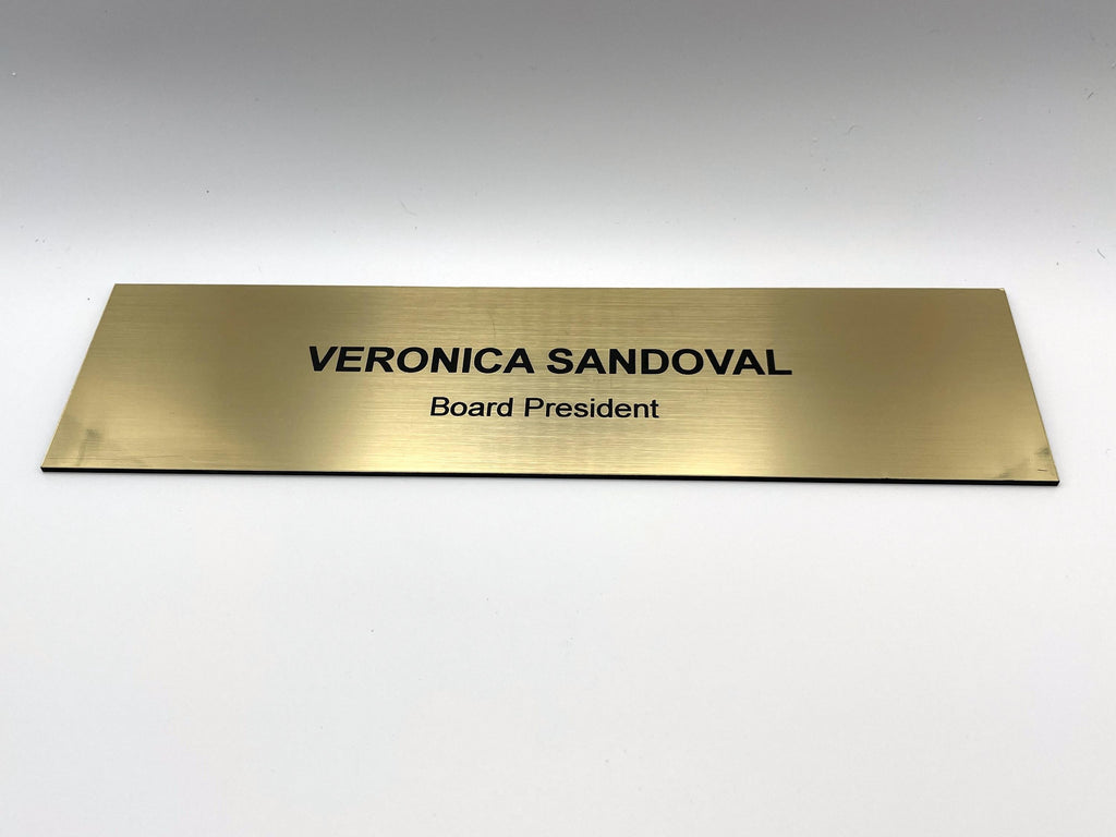 A rectangular gold-toned nameplate with a brushed metal-look finish. The text VERONICA SANDOVAL is engraved in bold black capital letters on the top line, with the title Board President in smaller black letters on the line below.