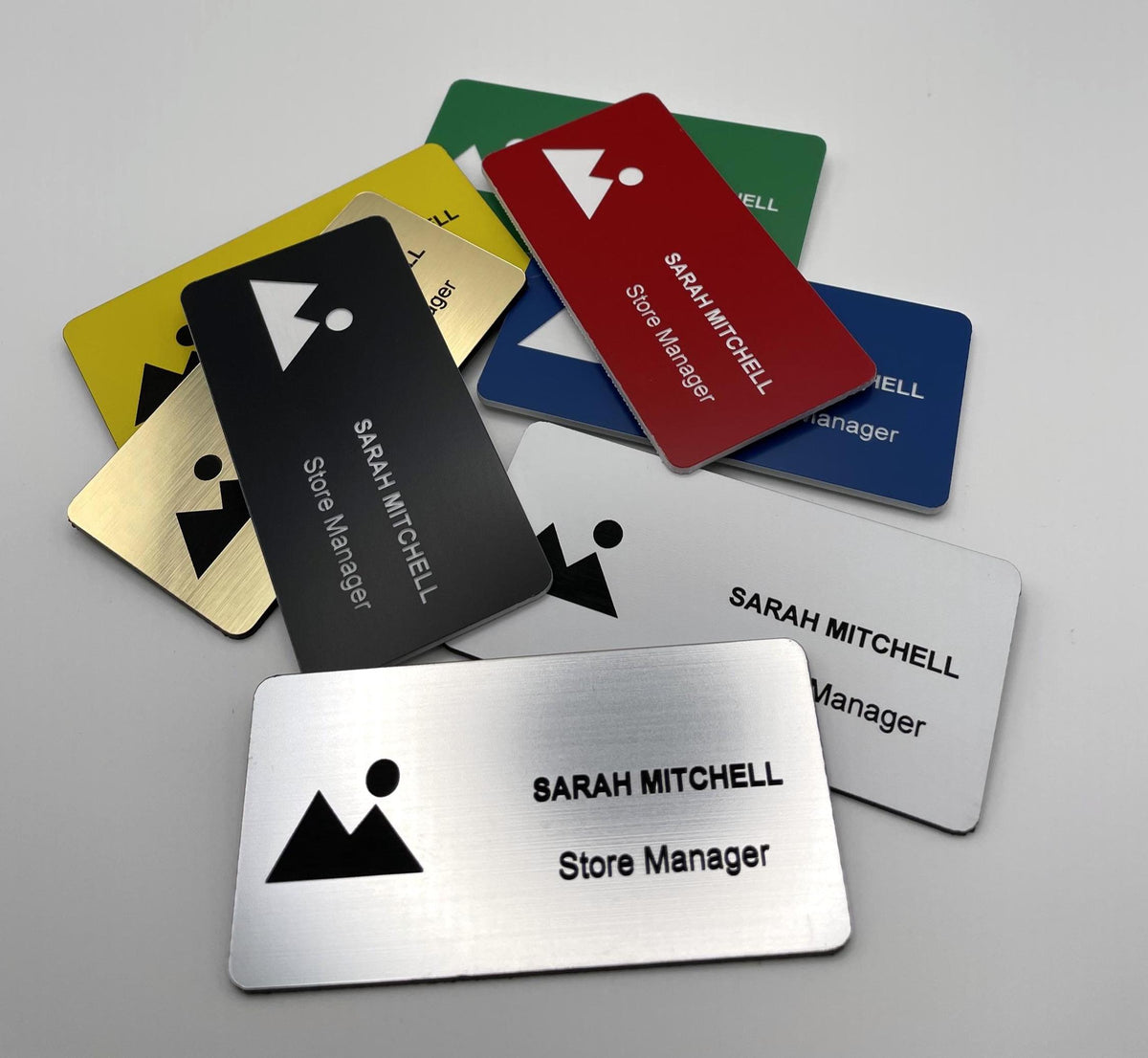 A collection of rectangular name badges in various colors including black, red, blue, green, yellow, silver, and gold. Each badge features a mountain-shaped logo with a circle above it, the name SARAH MITCHELL, and the title Store Manager.