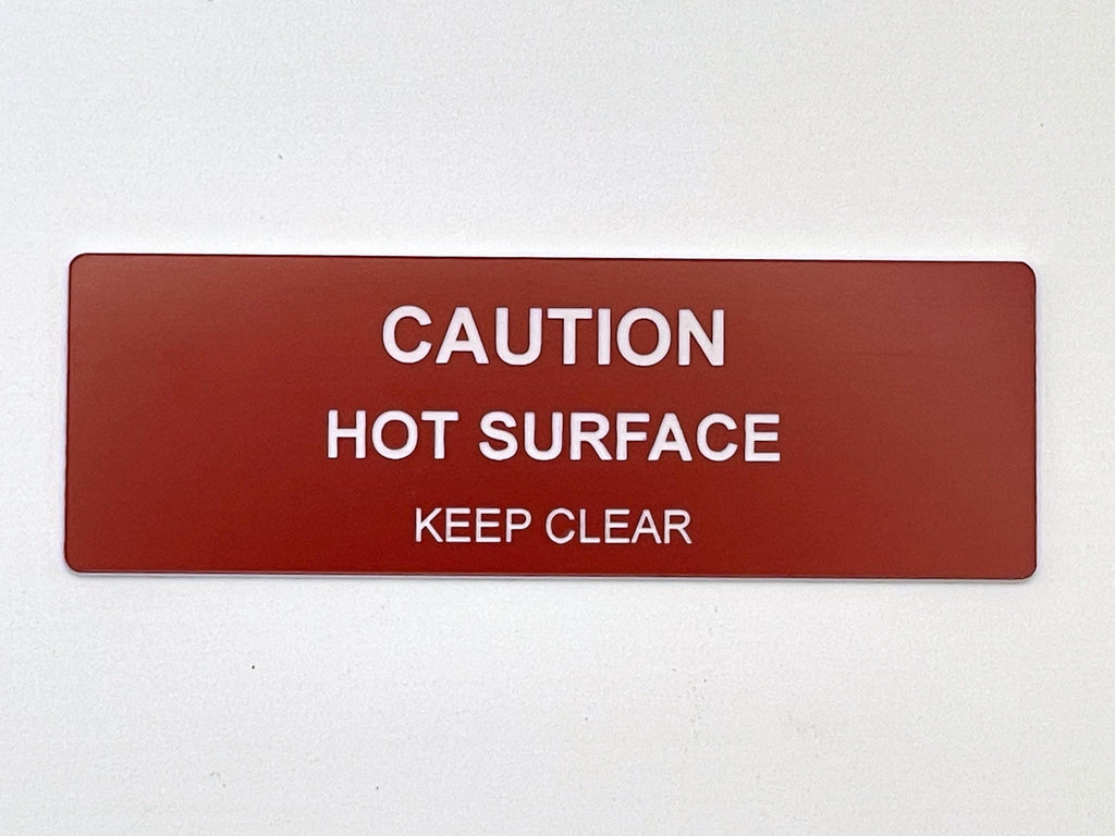 A rectangular red safety sign with rounded corners featuring white text that reads CAUTION HOT SURFACE KEEP CLEAR. The sign is designed for clear visibility and provides a warning message in a bold, sans-serif font style.