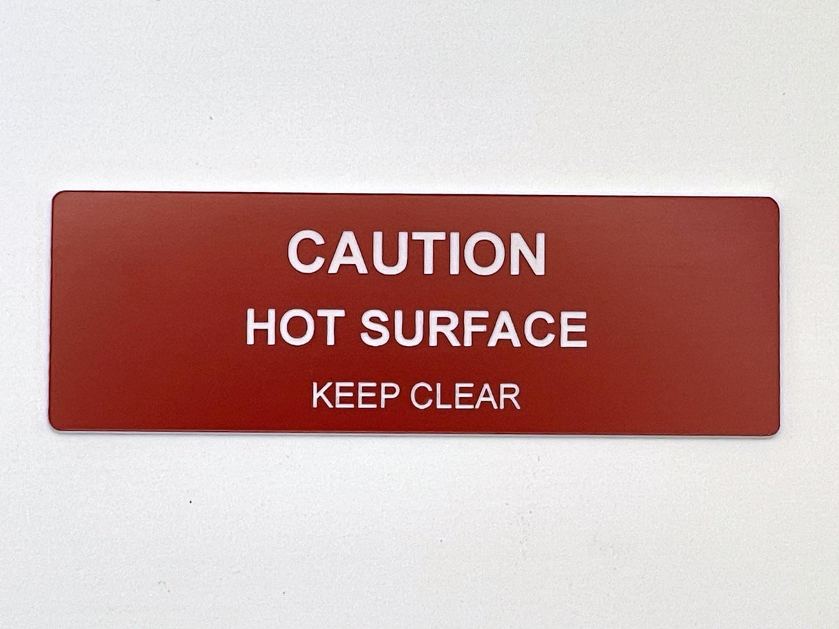 A rectangular red safety sign with rounded corners featuring white text that reads CAUTION HOT SURFACE KEEP CLEAR. The sign is designed for clear visibility and provides a warning message in a bold, sans-serif font style.