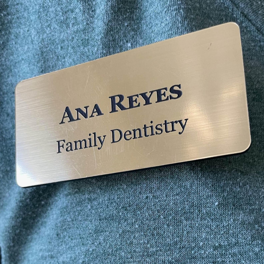 A rectangular brushed gold name badge with rounded corners. The text ANA REYES is engraved in capital letters on the top line, followed by the text Family Dentistry on the second line.