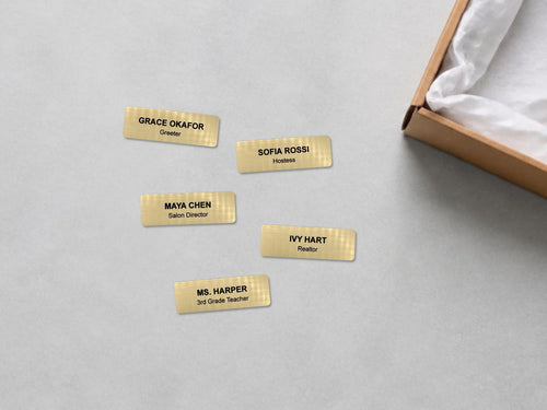 Five rectangular gold-toned name badges with a brushed metal-look texture. Each badge features black engraved text: Grace Okafor Greeter, Sofia Rossi Hostess, Maya Chen Salon Director, Ivy Hart Realtor, and Ms. Harper 3rd Grade Teacher.