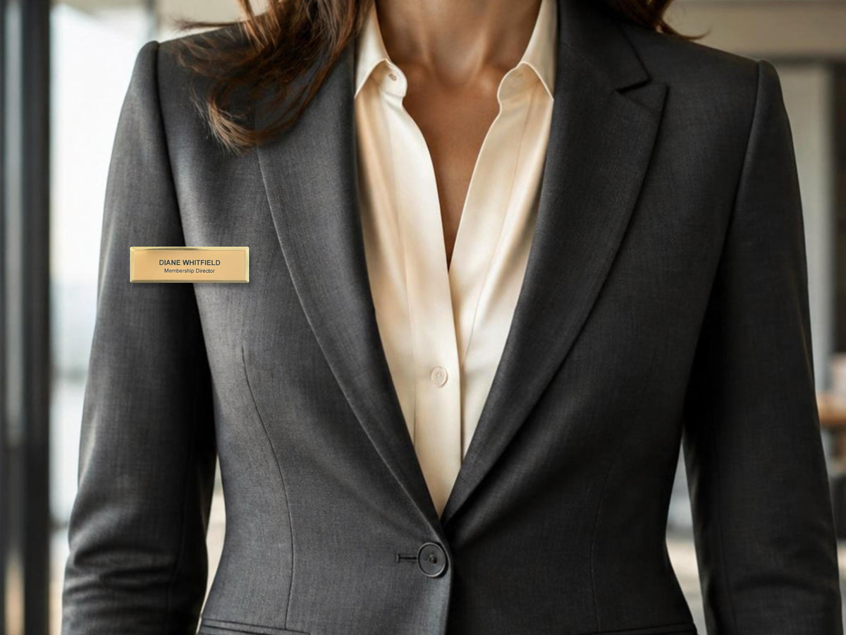 A rectangular gold-toned name badge pinned to a dark gray blazer. The badge features the text "DIANE WHITFIELD" on the top line and "Membership Director" on the bottom line in black lettering. The blazer is worn over a cream-colored shirt.
