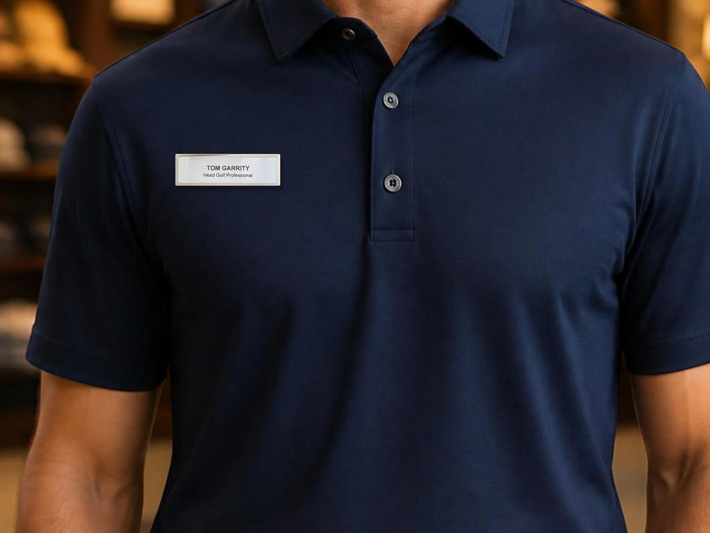 A navy blue short-sleeved polo shirt with a collar and a three-button placket. A rectangular silver name tag is pinned to the chest, featuring the text "TOM GARRITY" on the top line and "Head Golf Professional" below it.