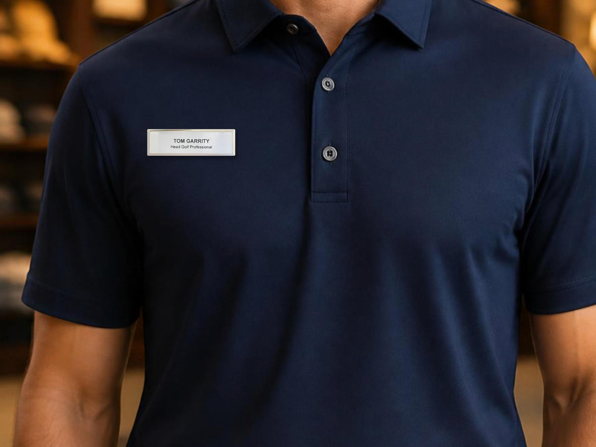 A navy blue short-sleeved polo shirt with a collar and a three-button placket. A rectangular silver name tag is pinned to the chest, featuring the text "TOM GARRITY" on the top line and "Head Golf Professional" below it.
