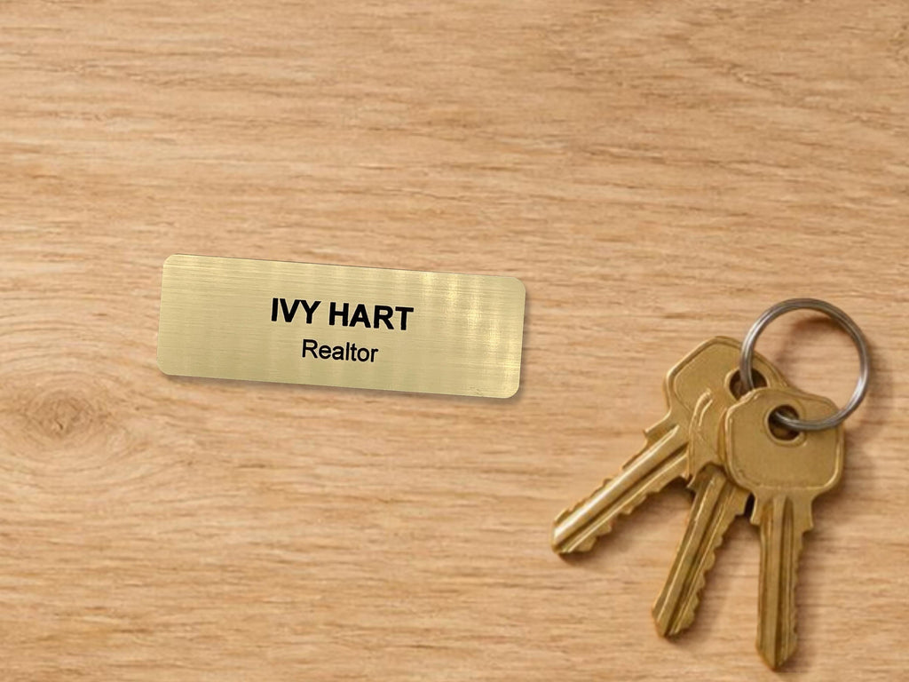 A rectangular gold-toned name tag with a brushed metal-look finish featuring the black engraved text IVY HART and Realtor, accompanied by a silver-toned metal keyring holding three brass-colored keys with a standard serrated edge design.