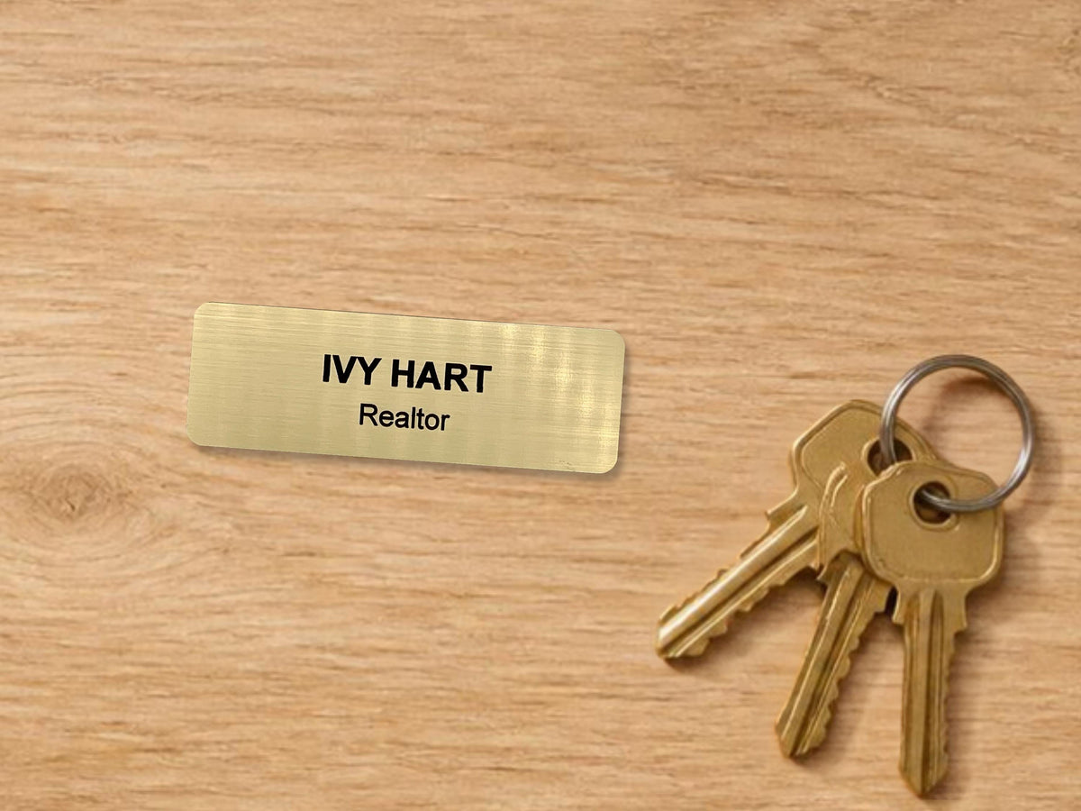 A rectangular gold-toned name tag with a brushed metal-look finish featuring the black engraved text IVY HART and Realtor, accompanied by a silver-toned metal keyring holding three brass-colored keys with a standard serrated edge design.