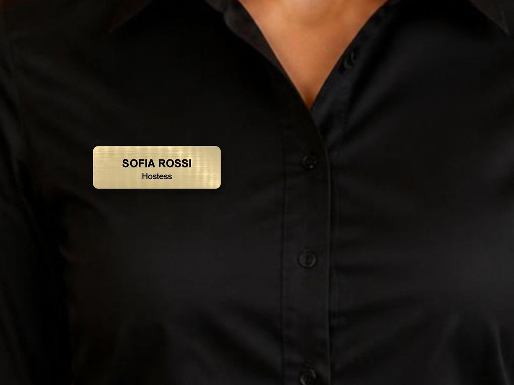 A rectangular gold-toned name badge with a brushed metal-look finish, featuring the black engraved text SOFIA ROSSI and the word Hostess underneath, pinned to a black button-down shirt with a collar and visible buttons.