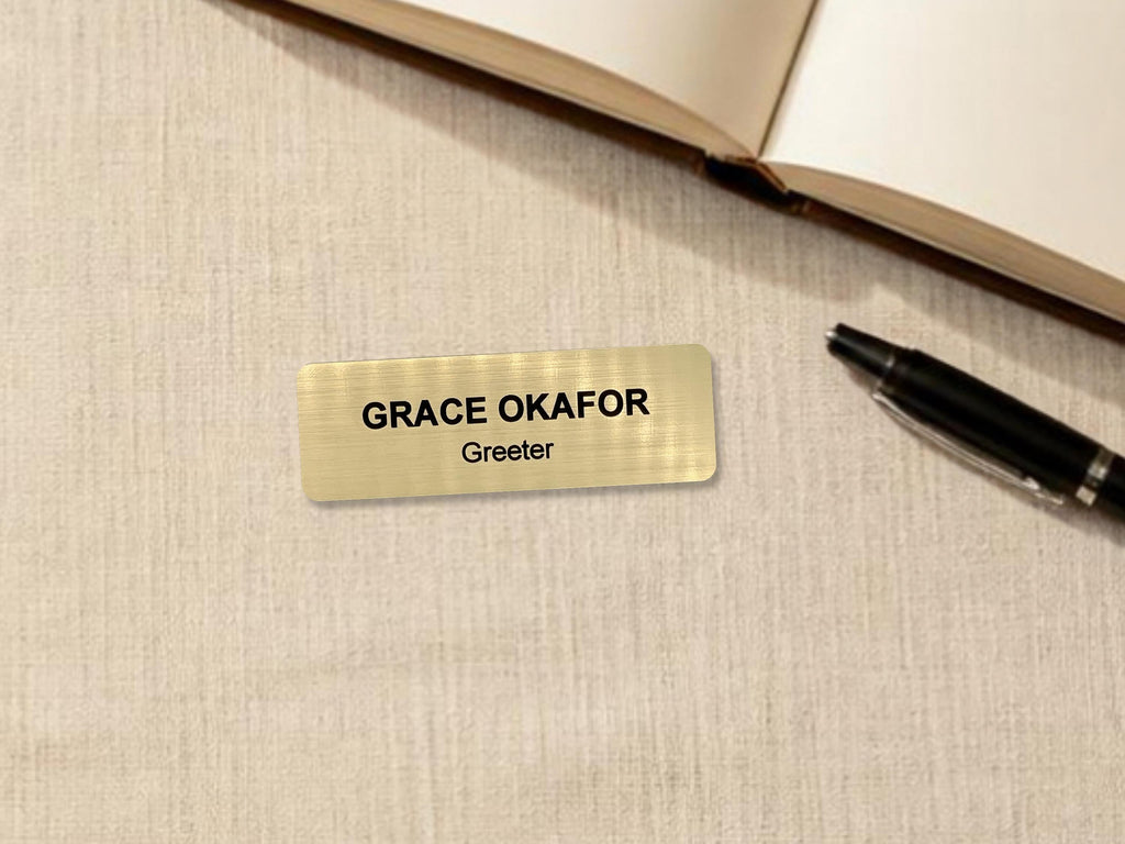 A rectangular gold-toned name badge with a brushed metal-look finish. The text "GRACE OKAFOR" is engraved in bold black capital letters on the top line, with the word "Greeter" in smaller black letters on the line below.