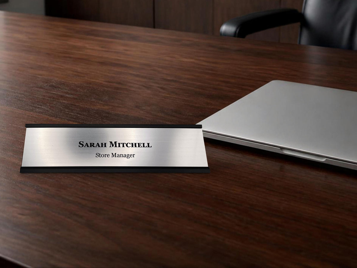 A rectangular desk nameplate featuring a brushed silver metal-look plate held in a black holder. The text Sarah Mitchell is engraved in a bold black serif font, with the title Store Manager in a smaller black font below it.