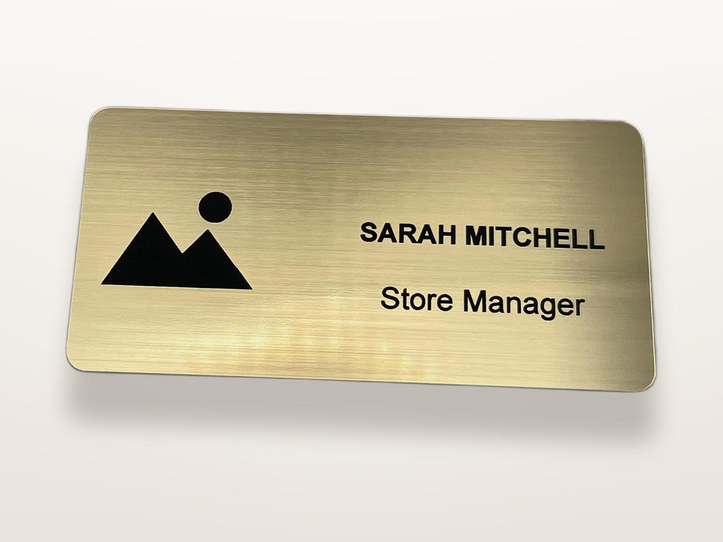 A rectangular brushed gold name badge with rounded corners. The left side features a black mountain and sun icon. The right side displays the text SARAH MITCHELL in uppercase letters, followed by the text Store Manager below it.