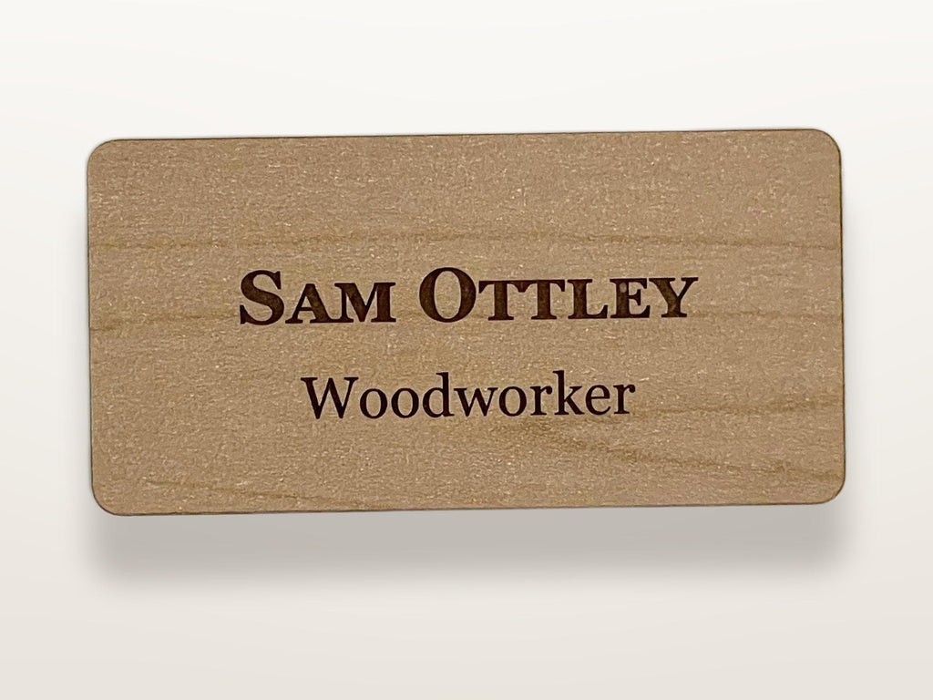 A rectangular light brown wooden name tag with rounded corners. The text "SAM OTTLEY" is engraved in a dark brown serif font on the top line, and the word "Woodworker" is engraved in the same dark brown font on the bottom line.