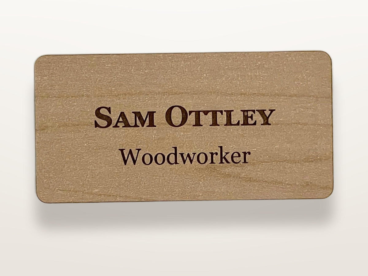 A rectangular light brown wooden name tag with rounded corners. The text "SAM OTTLEY" is engraved in a dark brown serif font on the top line, and the word "Woodworker" is engraved in the same dark brown font on the bottom line.