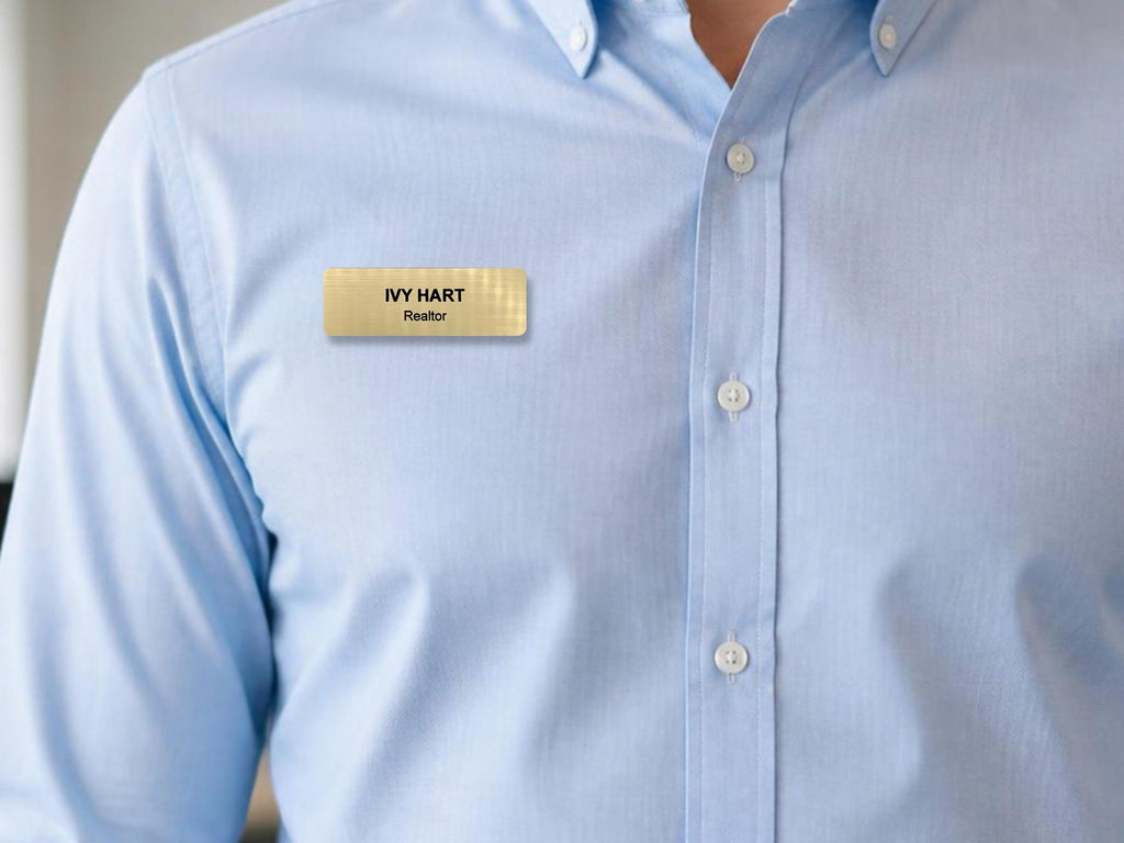 A rectangular gold-toned name badge with a subtle grid texture, featuring the black engraved text IVY HART and the title Realtor below it, pinned to a light blue button-down shirt with a classic collar and white buttons.