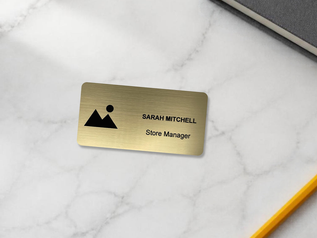 A rectangular brushed gold name badge featuring a black mountain and sun icon on the left side. The text SARAH MITCHELL is engraved in black capital letters on the top line, with the words Store Manager engraved below in black.
