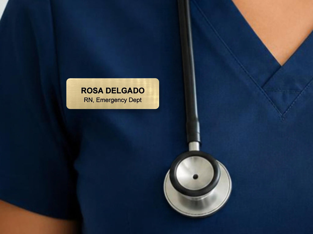 A rectangular gold-toned name badge with black text reading ROSA DELGADO, RN, Emergency Dept, pinned to a dark navy blue V-neck scrub top next to a black and silver stethoscope with a circular chest piece resting on the fabric.