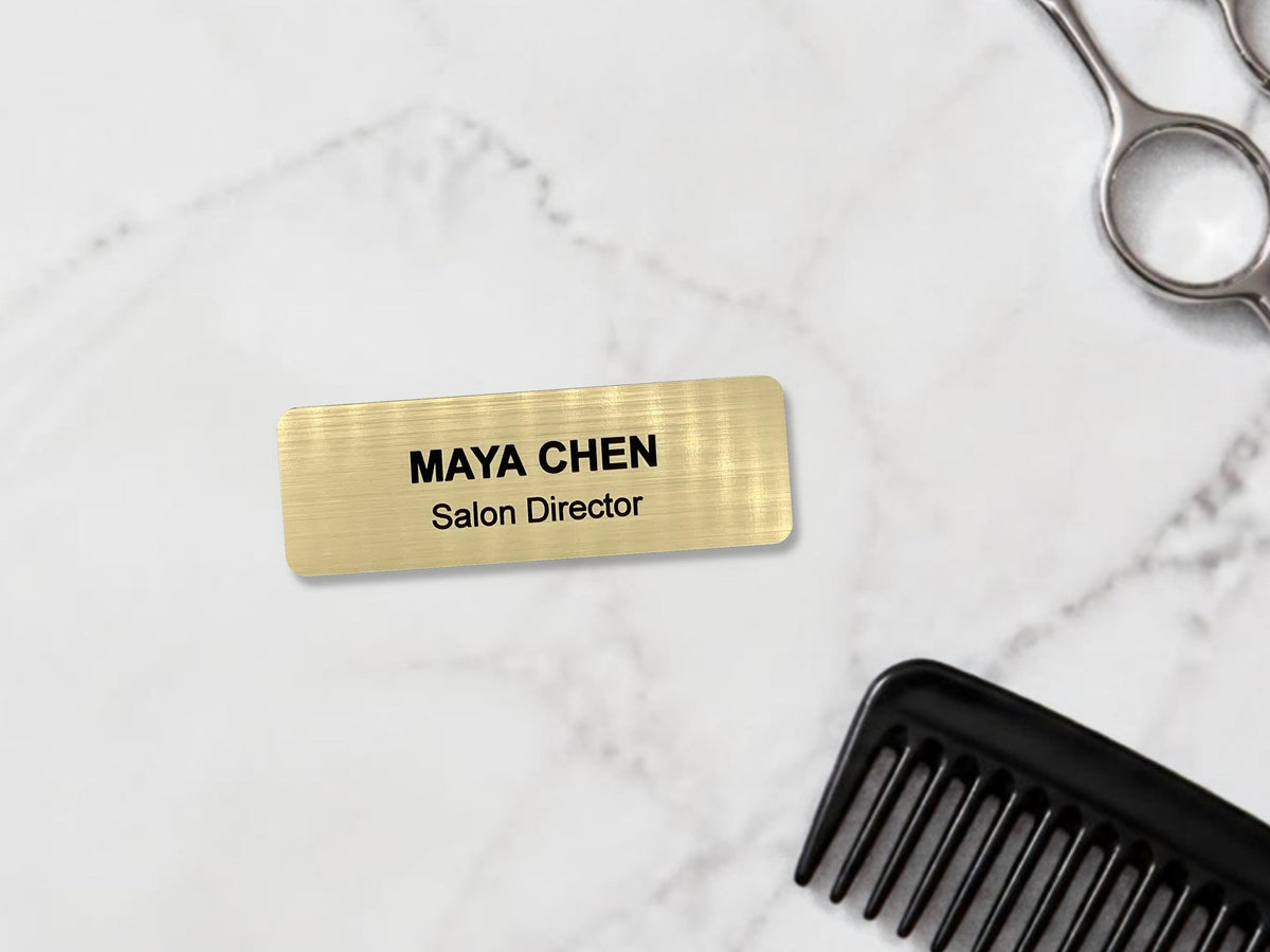 A rectangular gold-toned name badge with a brushed metal-look finish. The text "MAYA CHEN" is engraved in bold black capital letters on the top line, with "Salon Director" in smaller black letters on the line below.