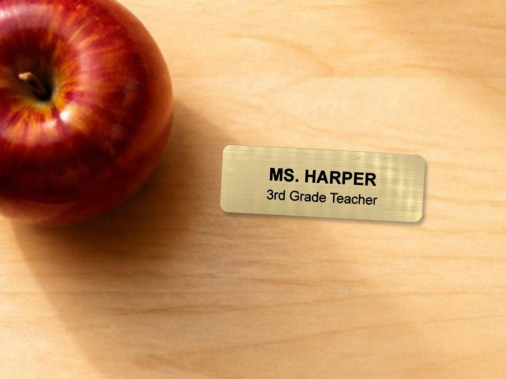 A rectangular gold-toned name tag with a brushed metal-look finish featuring the black engraved text MS. HARPER and 3rd Grade Teacher, positioned next to a shiny red apple with visible yellow highlights and a brown stem.