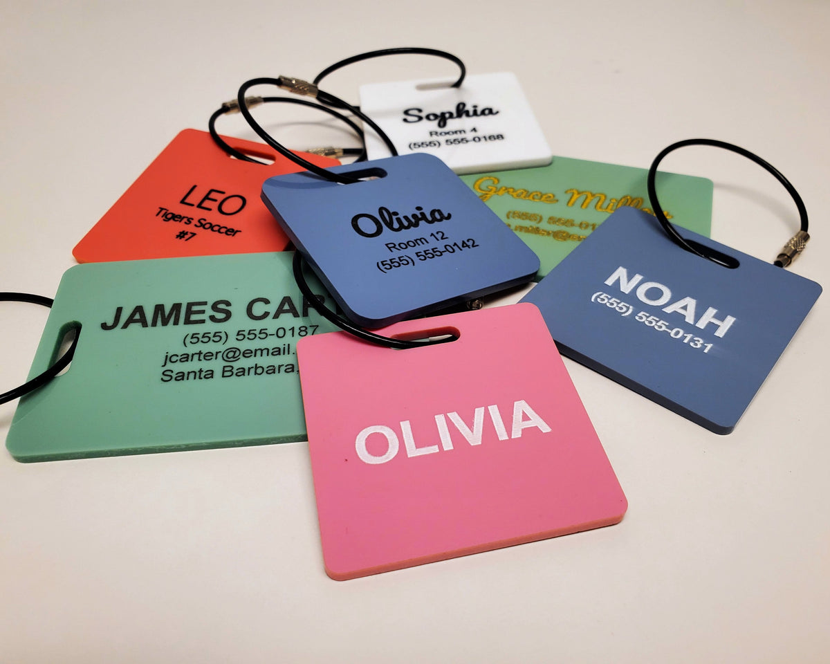 A pile of engraved pastel bag tags with black wire loops: OLIVIA in white on pink; LEO, Tigers Soccer #7 in black on coral; NOAH and a phone number in white on slate blue; Olivia in black script with Room 12 and a phone number on slate blue; Sophia with Room 4 and a phone number on glossy white; JAMES CARTER with a phone, email and city in black on sage green; and Grace Miller in gold on sage green.