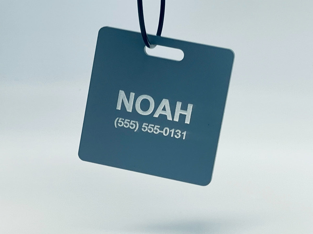 Square slate blue bag tag hanging from its wire loop, engraved NOAH and (555) 555-0131 in white.