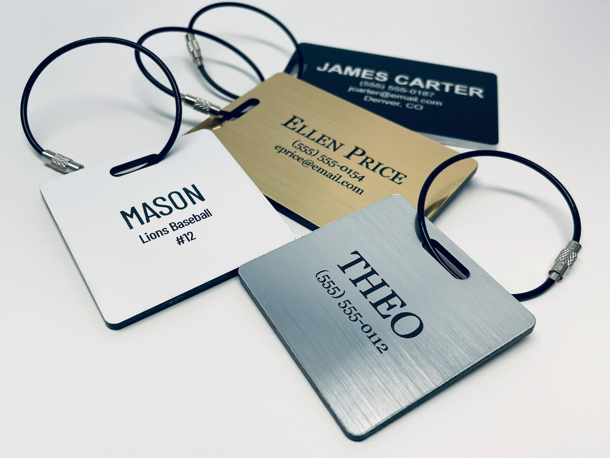 Bag tags in the classic colors: MASON, Lions Baseball #12 on white; ELLEN PRICE on brushed gold; THEO on brushed silver; and JAMES CARTER on black, each with a black wire loop.