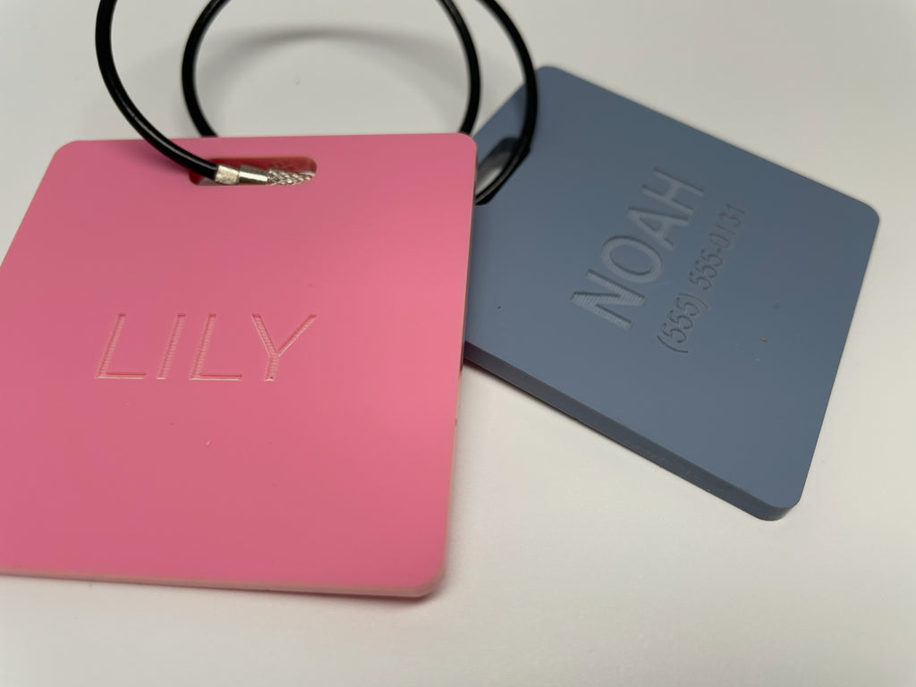 Two pastel bag tags with frosted, unpainted lettering: LILY on pink and NOAH with a phone number on slate blue. The frosted letters are subtle, tone on tone.