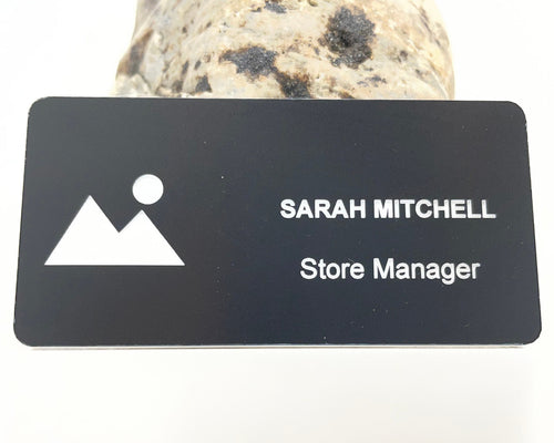 A matte black engraved name badge with a white mountain and sun logo and SARAH MITCHELL, Store Manager, in white, standing in front of a speckled stone.
