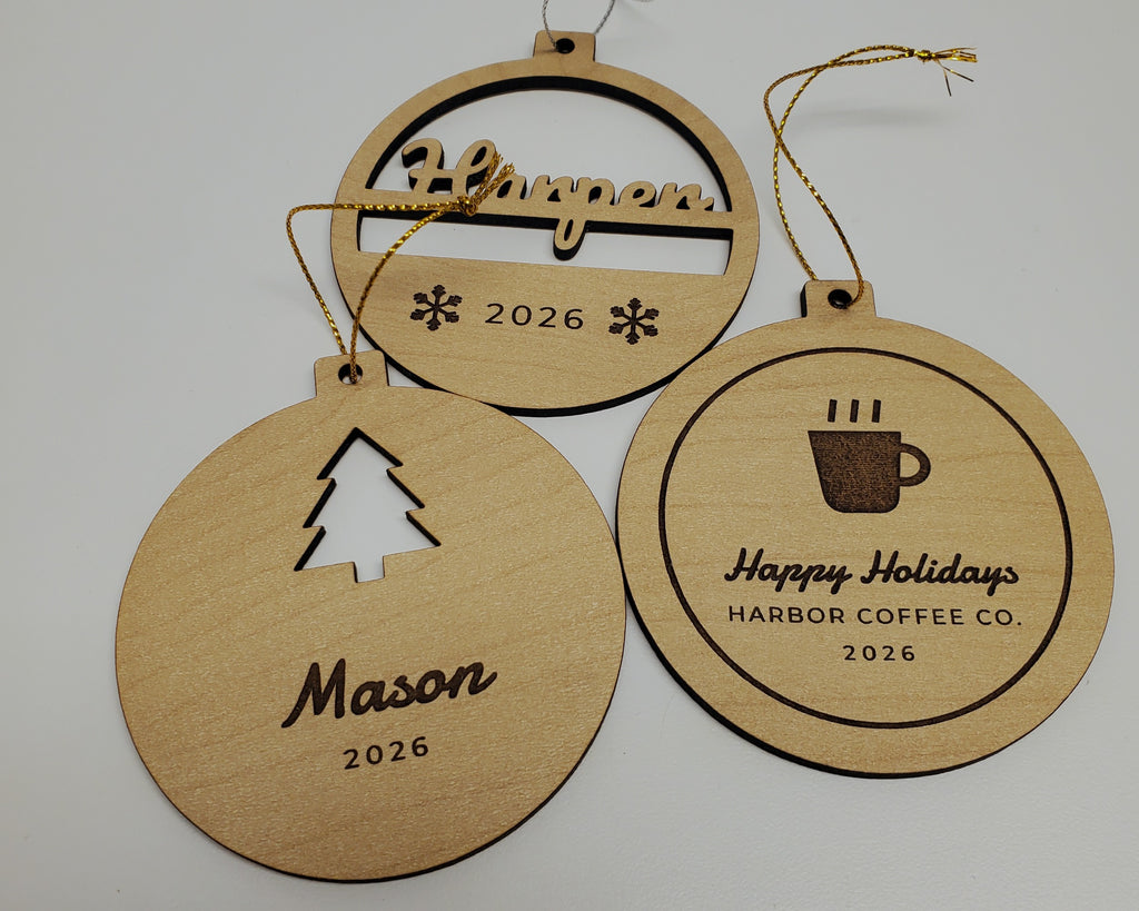 Three personalized Christmas ornaments in maple finish on metallic gold and silver string: Harper cut through in script above 2026 and two engraved snowflakes; Mason with a tree cut out above the name and 2026; and a Happy Holidays, HARBOR COFFEE CO., 2026 ornament with a coffee cup logo.