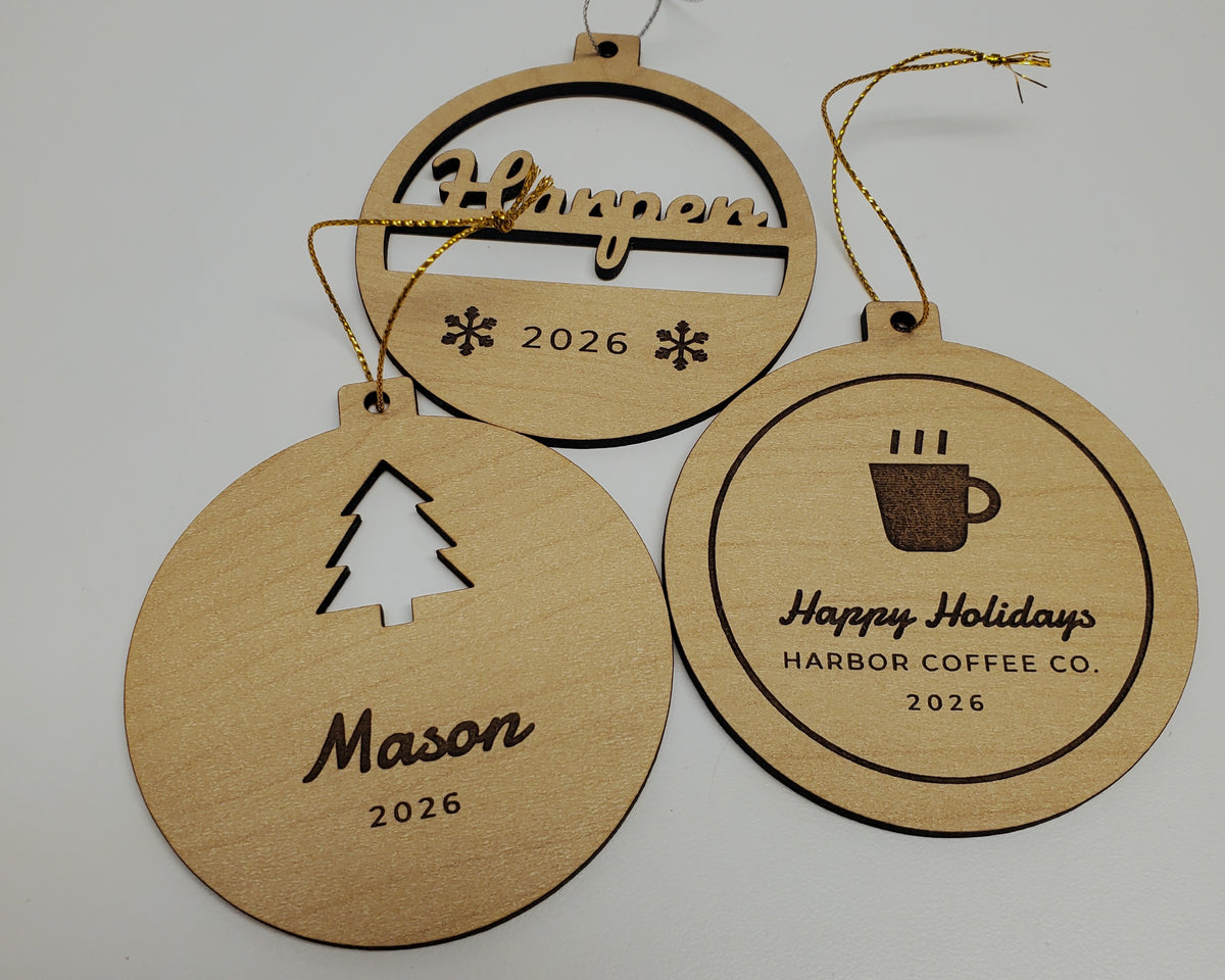 Three personalized Christmas ornaments in maple finish on metallic gold and silver string: Harper cut through in script above 2026 and two engraved snowflakes; Mason with a tree cut out above the name and 2026; and a Happy Holidays, HARBOR COFFEE CO., 2026 ornament with a coffee cup logo.