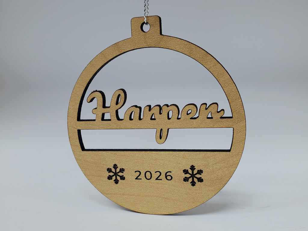 A Harper Christmas ornament in maple finish hanging from metallic silver string: the name cut through in script, standing on a crossbar, with 2026 and two snowflakes engraved below.