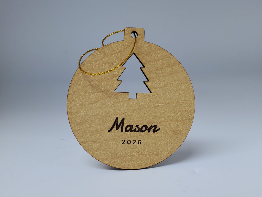 A Mason Christmas ornament in maple finish standing upright: a tree cut out of the round, with Mason in script and 2026 engraved below, and its gold string looped over the top.