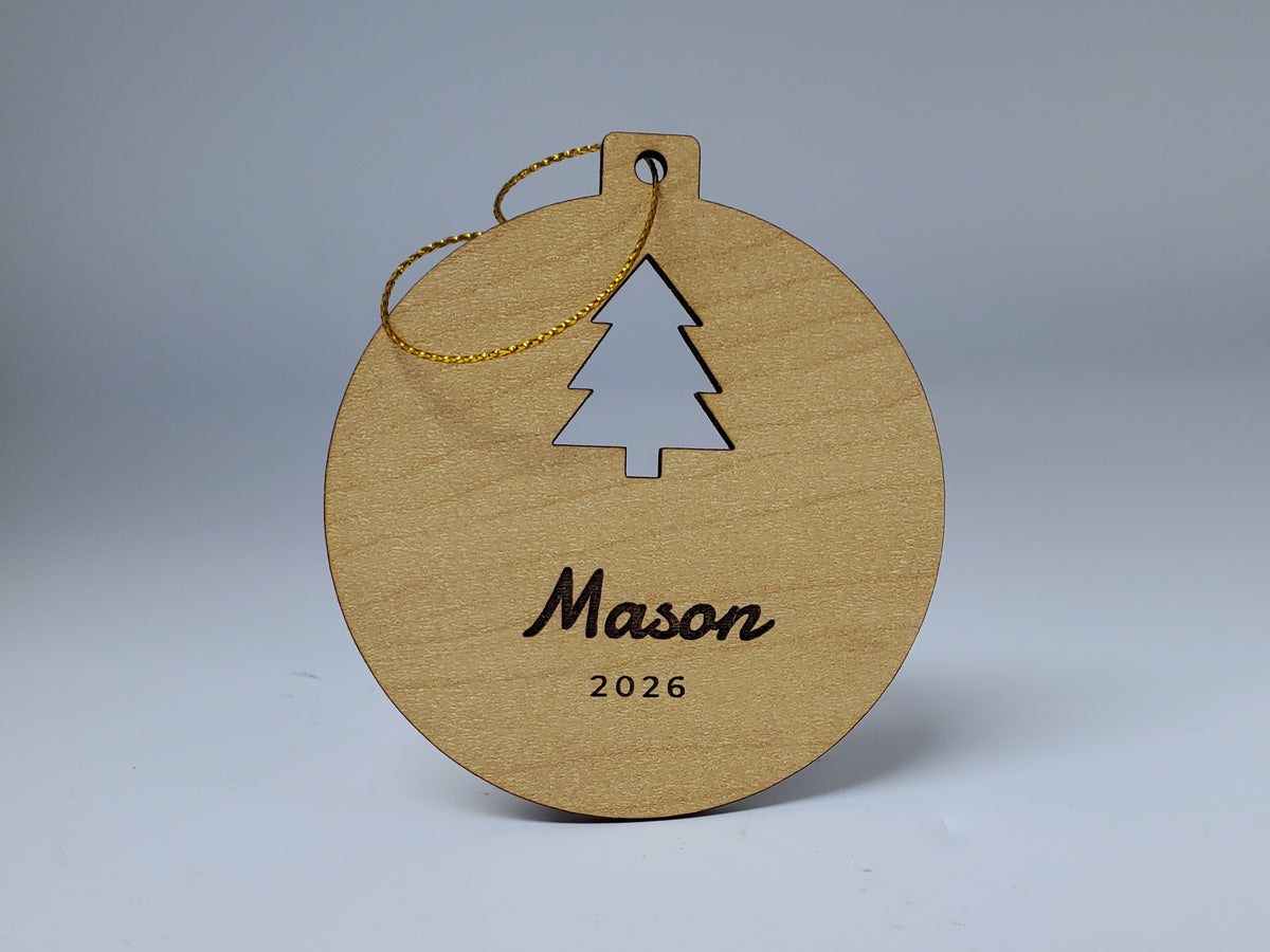 A Mason Christmas ornament in maple finish standing upright: a tree cut out of the round, with Mason in script and 2026 engraved below, and its gold string looped over the top.