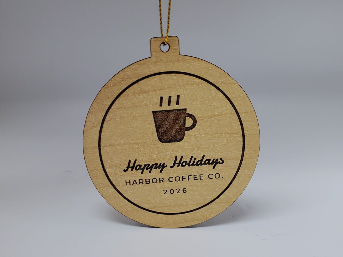 A business Christmas ornament in maple finish hanging from metallic gold string: an engraved coffee cup logo, Happy Holidays in script, HARBOR COFFEE CO. and 2026, inside an engraved border ring.