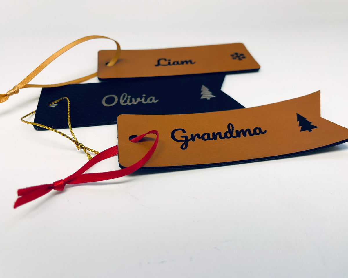 Three Christmas stocking name tags lying together: Liam on rawhide leatherette with a snowflake and gold ribbon, Olivia in gold on black leatherette with a tree, and Grandma on a rawhide flag tag with a tree and red ribbon.
