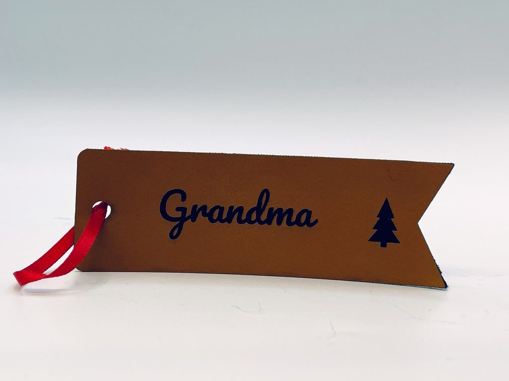 Rawhide leatherette flag stocking tag standing on a table: Grandma in script and a small tree, with a red ribbon through the hole.