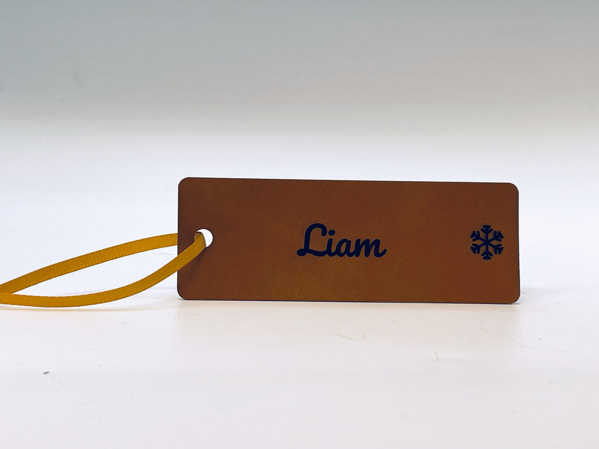 Rawhide leatherette stocking tag with rounded corners: Liam in script and a snowflake, with a gold ribbon loop.