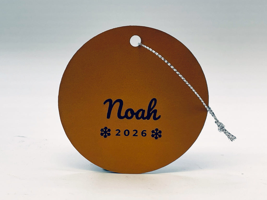 Round rawhide leatherette stocking tag: Noah in script over 2026 between two snowflakes, with silver string.