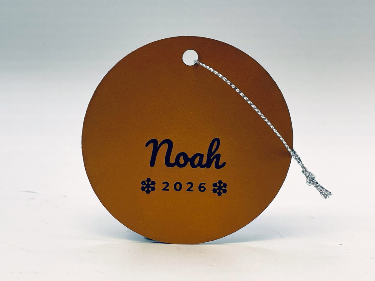 Round rawhide leatherette stocking tag: Noah in script over 2026 between two snowflakes, with silver string.
