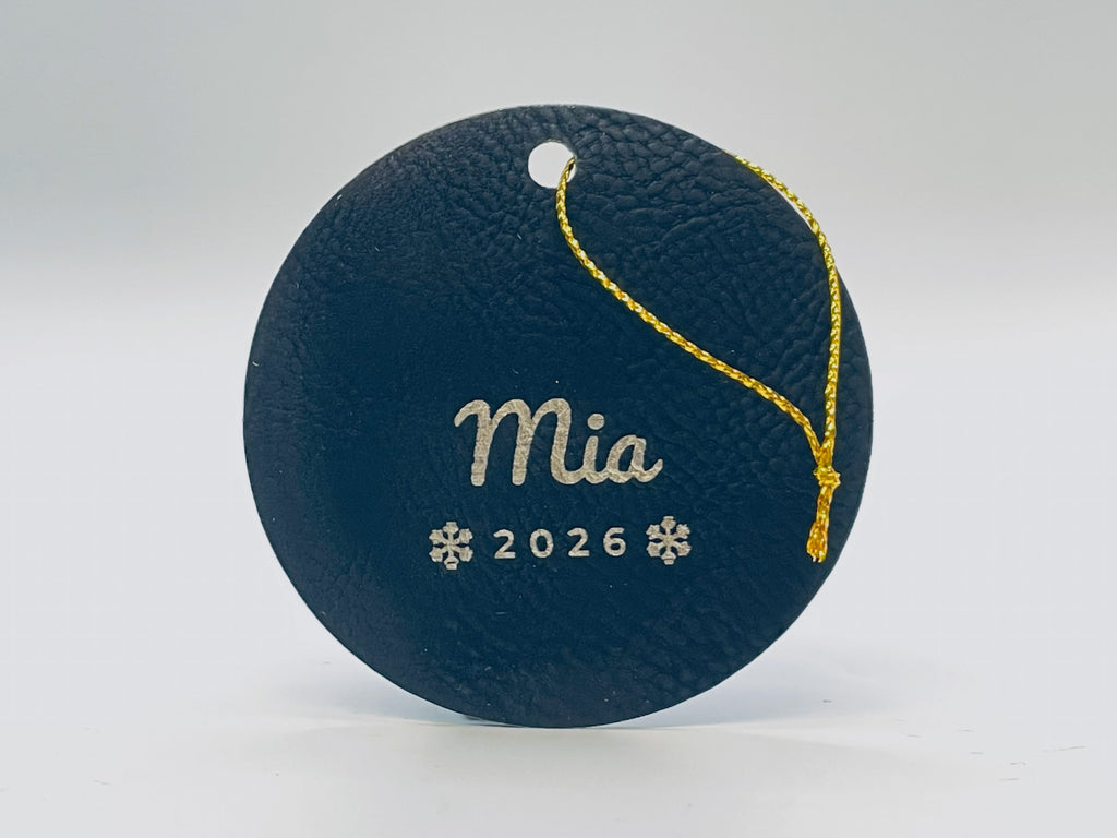 Round black leatherette stocking tag engraved in gold: Mia in script over 2026 between two snowflakes, with gold string.