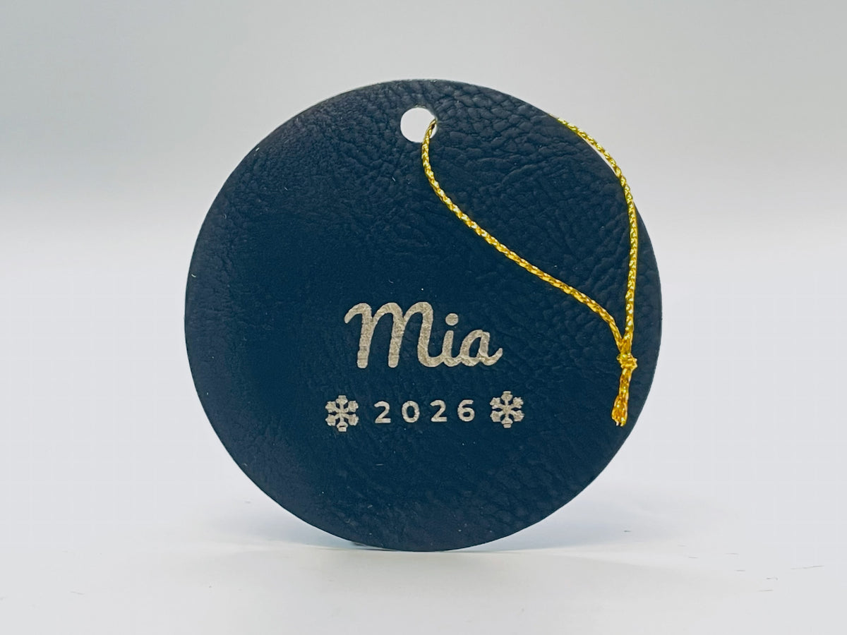 Round black leatherette stocking tag engraved in gold: Mia in script over 2026 between two snowflakes, with gold string.