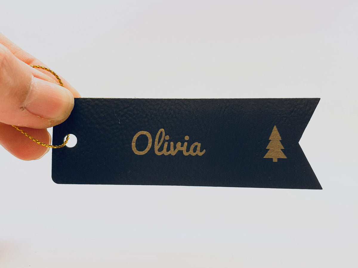 A hand holding the black and gold Olivia flag tag by its gold string, for size.