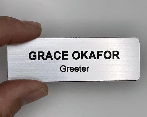 A rectangular silver-toned name badge with a brushed metal-look finish. The badge features the black engraved text GRACE OKAFOR on the top line and Greeter on the bottom line. The edges of the name tag are rounded and have a black border.
