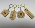 Four engraved keychains in maple finish lying in a row, each on a split ring and chain: a house reading HOME Sweet HOME, EMILY CARTER and a phone number; a motel tag reading SUNSET INN, 12, Good vibes only; a round HARBOR COFFEE CO. logo keychain; and a bar reading MARTINEZ ELECTRIC with a license and phone line. Silver and gold rings.