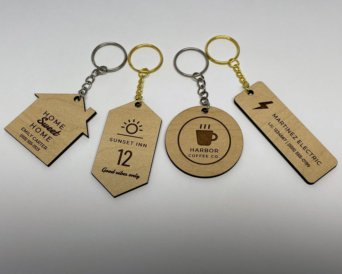 Four engraved keychains in maple finish lying in a row, each on a split ring and chain: a house reading HOME Sweet HOME, EMILY CARTER and a phone number; a motel tag reading SUNSET INN, 12, Good vibes only; a round HARBOR COFFEE CO. logo keychain; and a bar reading MARTINEZ ELECTRIC with a license and phone line. Silver and gold rings.