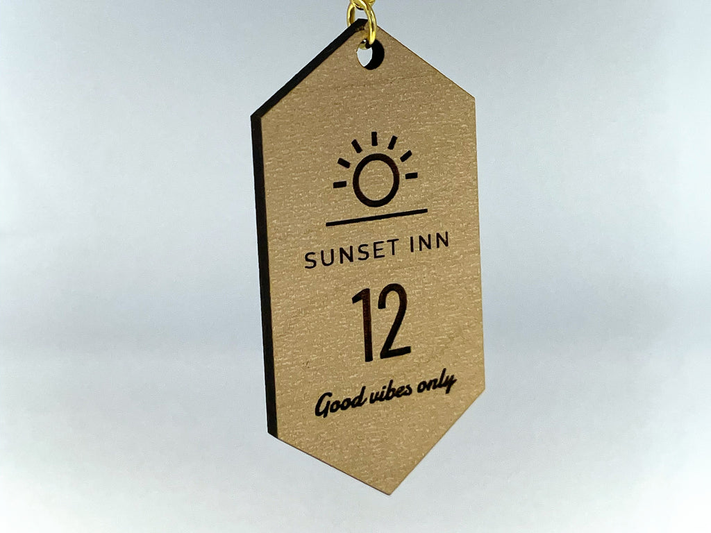 A motel tag keychain in maple finish hanging from its gold chain, engraved with a sun logo, SUNSET INN, the number 12 and Good vibes only.